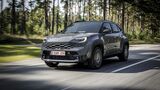 Grauer Toyota Yaris Cross Hybrid 130 GR Sport in schräger Frontansicht auf einer Straße mit Wald im Hintergrund.