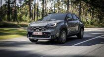 Grauer Toyota Yaris Cross Hybrid 130 GR Sport in schräger Frontansicht auf einer Straße mit Wald im Hintergrund.