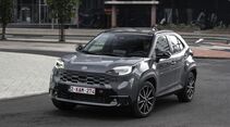 Grauer Toyota Yaris Cross Hybrid 130 GR Sport in schräger Frontansicht auf einer städtischen Straße mit Gebäuden im Hintergrund.