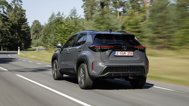 Grauer Toyota Yaris Cross Hybrid 130 GR Sport in schräger Heckansicht während der Fahrt auf einer Landstraße.