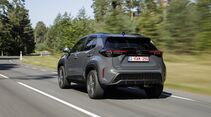 Grauer Toyota Yaris Cross Hybrid 130 GR Sport in schräger Heckansicht während der Fahrt auf einer Landstraße.
