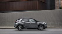 Grauer Toyota Yaris Cross Hybrid 130 GR Sport mit schwarzem Dach in Seitenansicht während der Fahrt vor einer Betonwand.