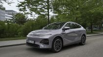 Grauer XPeng G6 Elektro-SUV fährt auf asphaltierter Straße durch städtische Umgebung mit Bäumen im Hintergrund.
