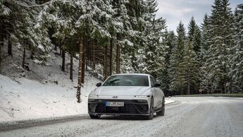 Hyundai Ioniq 6 frontal auf verschneiter Landstraße im Wald, Winterwetter, Fahrbahn mit Schneeresten.