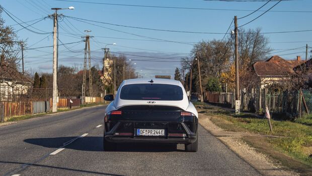 Hyundai Ioniq 6 von hinten auf Landstraße, Leitungen und Häuser am Straßenrand, klare Wetterlage.
