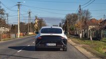Hyundai Ioniq 6 von hinten auf Landstraße, Leitungen und Häuser am Straßenrand, klare Wetterlage.