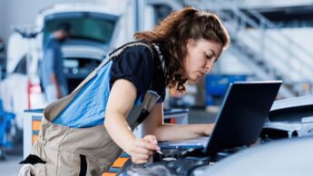 Kfz-Mechanikerin liest mit Laptop Fahrzeugdaten aus dem offenen Motorraum eines Autos in einer Werkstatt aus