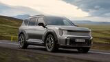 Kia EV9 GT als graues Elektro-SUV bei der Fahrt auf einer Landstrasse vor einer weitlaeufigen Berglandschaft.