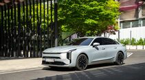 Kia K8 in Front- und Seitenansicht vor modernem Gebäude mit Grünanlagen.