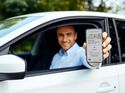 Mann im Auto hält ein Smartphone mit digitalem Fahrzeugschein und Führerschein-App aus dem Fenster in die Kamera.