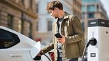 Mann lädt ein weißes Elektroauto an einer öffentlichen Ladesäule und hält ein Smartphone in der Hand. 