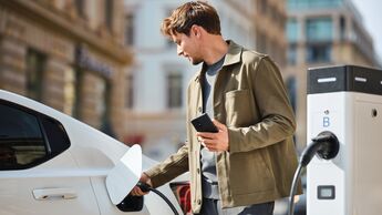 Mann lädt ein weißes Elektroauto an einer öffentlichen Ladesäule und hält ein Smartphone in der Hand. 