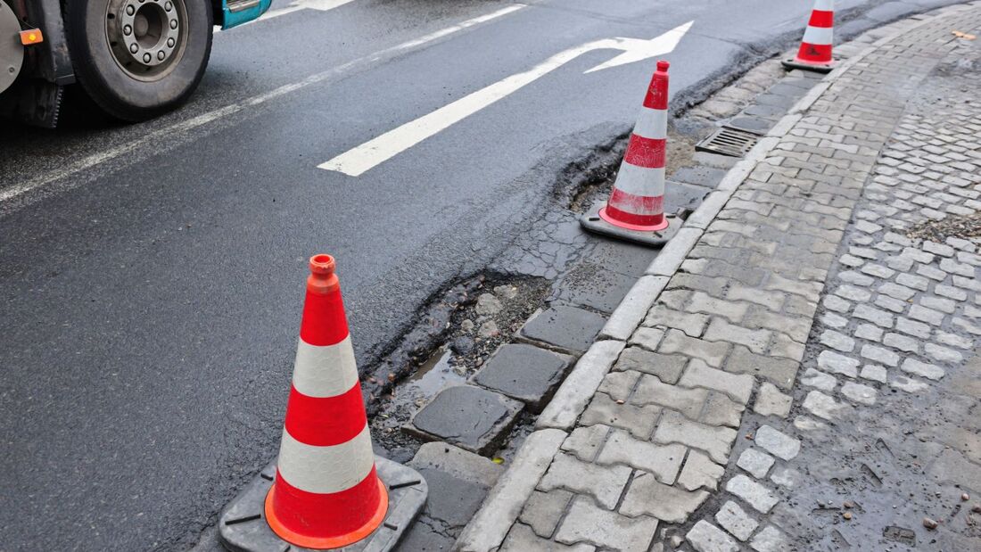 Mit Leitkegeln abgesicherte Schlaglöcher am rechten Fahrbahnrand einer innerstädtischen Straße neben fließendem Verkehr.