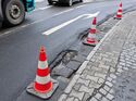 Mit Leitkegeln abgesicherte Schlaglöcher am rechten Fahrbahnrand einer innerstädtischen Straße neben fließendem Verkehr.
