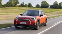 Orangefarbener Mini Aceman SE in Front-Dreiviertelansicht während der Fahrt auf einer Landstraße.