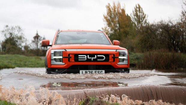 Oranger BYD Shark DMO bei einer Wasserdurchfahrt, frontal aufgenommen, mit aufgewirbeltem Wasser vor dem Fahrzeug.