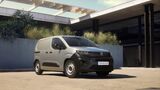 Peugeot E-Partner Active als grauer elektrischer Kastenwagen in Dreiviertelansicht von vorne, geparkt vor einem modernen Gebäude.