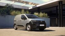 Peugeot E-Partner Active als grauer elektrischer Kastenwagen in Dreiviertelansicht von vorne, geparkt vor einem modernen Gebäude.