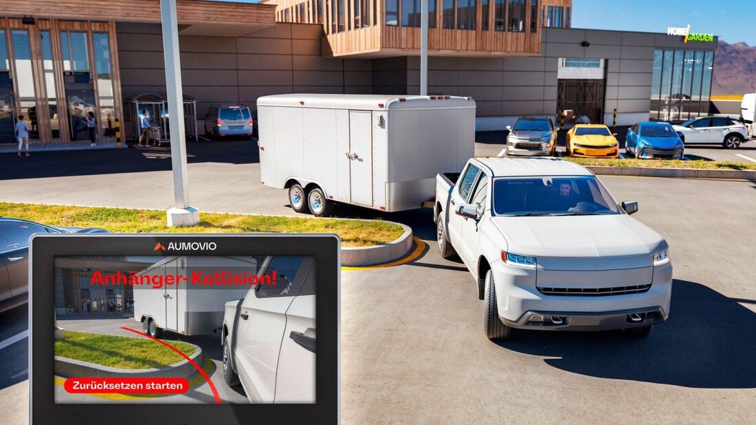 Pickup mit anhaenger beim rangieren; display zeigt aumovio warnung „anhaenger-kollision“ auf einem parkplatz vor gebaeude.