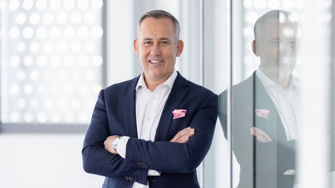 Portrait von Anthony Bandmann im Business-Outfit, stehend mit verschränkten Armen vor Glaswand in einem Bürogebäude.