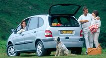 Renault Scenic in Silber mit geoeffneter Heckklappe auf Wiese; Personen und Picknickzubehoer stehen neben dem Fahrzeug, Hund davor.
