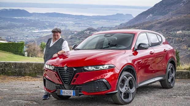 Roter Alfa Romeo Tonale in schräger Frontansicht, eine Person steht neben dem Fahrzeug, Landschaft und Tal im Hintergrund.