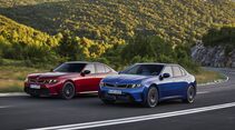 Roter BMW 3er und blauer BMW i3 fahren nebeneinander auf einer Straße durch eine bewaldete Berglandschaft.