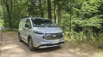 Schräg von vorn fotografierter Ford E-Transit Custom AWD Trail auf einem Waldparkplatz.
