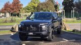 Schwarzer Mitsubishi L200 mit Offroad-Bereifung, Schnorchel und schwarzem Kühlergrill in schräger Frontansicht auf einem Parkplatz.
