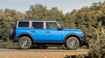 Seitenansicht eines blauen Ford Bronco während der Fahrt auf befestigter Straße mit Bäumen im Hintergrund.