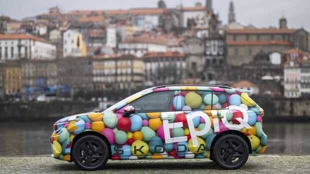 Seitliche Ansicht des Skoda Epiq Elektro-SUV mit bunter Tarnfolie vor historischer Stadtkulisse am Wasser.