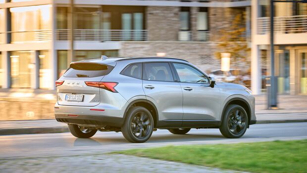 Silberner GWM Haval H6 in Heckansicht während der Fahrt vor modernen Gebäuden.