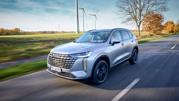 Silberner GWM Haval H6 während der Fahrt auf einer Landstraße vor Windrädern.