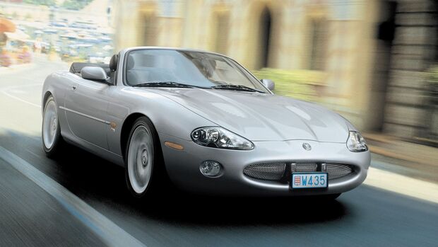 Silberner Jaguar XK8 Cabrio in Frontansicht waehrend der Fahrt auf Strasse, Verdeck geoeffnet, Hintergrund mit Bewegungsunschärfe.