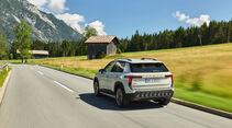 Skoda Epiq von hinten in Fahrt auf einer Landstraße vor einer Holzscheune in den Alpen.