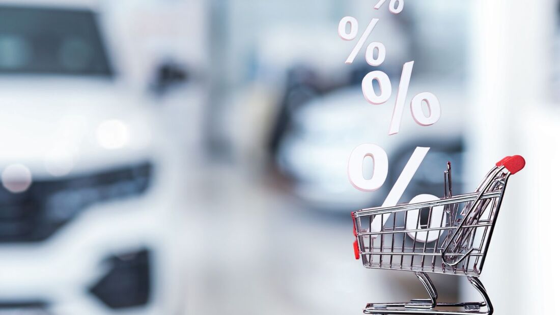 Symbolisches Einkaufswagensymbol mit Prozentzeichen vor unscharfen Elektrofahrzeugen, Darstellung von Preisentwicklung im Gebrauchtwagenmarkt.