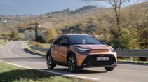 Toyota Aygo X Hybrid fährt auf einer Landstraße, Frontansicht in Cinnamon-Lackierung.