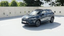 Toyota Corolla Cross in Grau, Front-Dreiviertelansicht, stehendes SUV auf Parkdeck vor Betonwand und Baeumen.