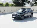 Toyota Corolla Cross in Grau, Front-Dreiviertelansicht, stehendes SUV auf Parkdeck vor Betonwand und Baeumen.