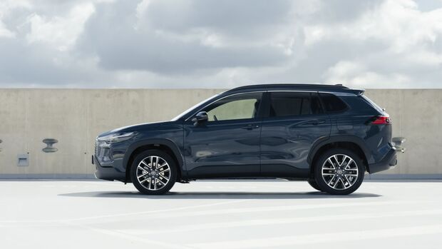 Toyota Corolla Cross in Grau, Seitenansicht, stehendes SUV auf Parkdeck vor Betonwand und bewolktem Himmel.