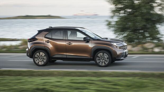 Toyota Yaris Cross Hybrid 130 in Bronze mit schwarzem Dach in Seitenansicht während der Fahrt auf einer Straße am Wasser.