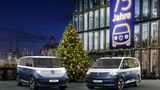 VW ID. Buzz und VW Multivan Sondermodelle vor einem Gebaeude mit Banner „75 Jahre“ und Weihnachtsbaum bei Nacht am Hafen.