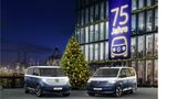 VW ID. Buzz und VW Multivan Sondermodelle vor einem Gebaeude mit Banner „75 Jahre“ und Weihnachtsbaum bei Nacht am Hafen.