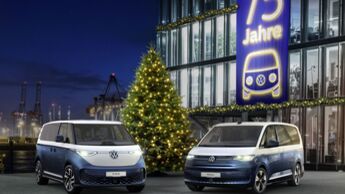 VW ID. Buzz und VW Multivan Sondermodelle vor einem Gebaeude mit Banner „75 Jahre“ und Weihnachtsbaum bei Nacht am Hafen.