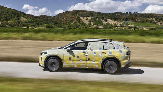 VW ID. Tiguan als getarnter Prototyp in gelb-grauer Folierung, Seitenansicht in voller Fahrt auf einer Landstraße vor hügeliger Landschaft