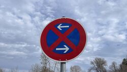 Verkehrsschild absolutes Halteverbot mit Richtungsangaben am Straßenrand vor bewölktem Himmel.
