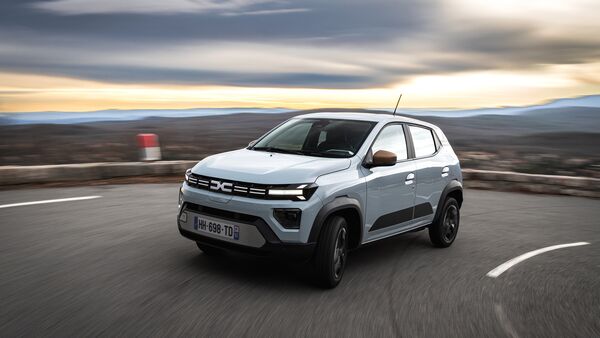 Weißer Dacia Spring Elektro-Kleinwagen fährt auf kurviger Straße in ländlicher Umgebung.