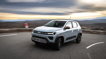 Weißer Dacia Spring Elektro-Kleinwagen fährt auf kurviger Straße in ländlicher Umgebung.