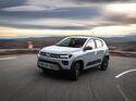 Weißer Dacia Spring Elektro-Kleinwagen fährt auf kurviger Straße in ländlicher Umgebung.