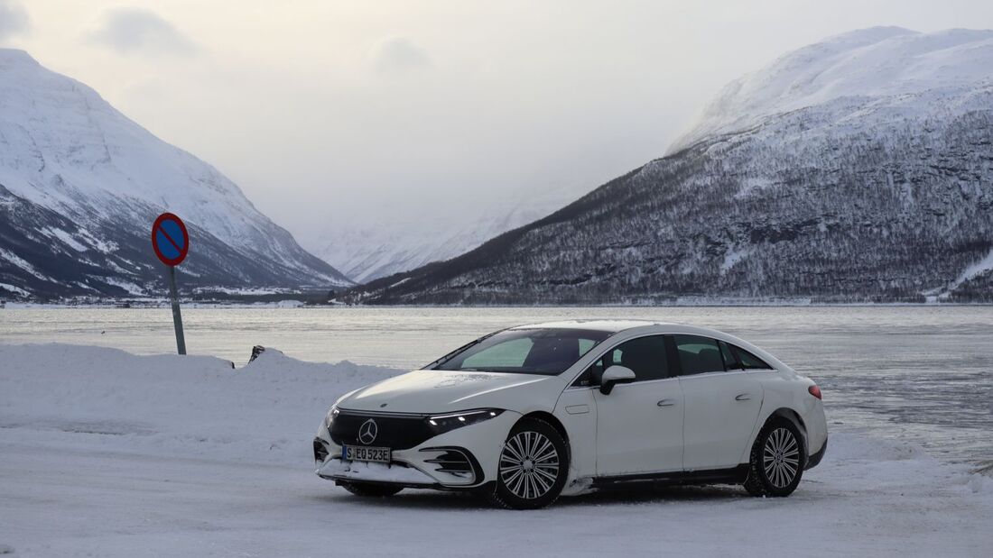 Weisses Elektroauto auf verschneiter Strasse vor winterlicher Berglandschaft und gefrorenem See.
